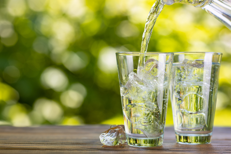 Zwei Gläser stehen auf einem Tisch, beide sind mit Eiswürfeln gefüllt, in ein Glas wird Wasser eingeschenkt.