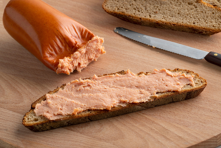 Auf einem Holzbrett liegt eine Teewurst neben einem Brot, das mit Teewurst bestrichen ist.