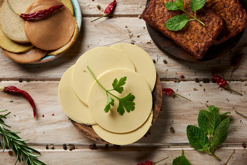 Blick aus der Vogelperspektive auf einige Scheiben veganen Käses, einige Tofu-Filets und einige Aufschnitte aus Tofu auf einem weißen, rustikalen Tisch