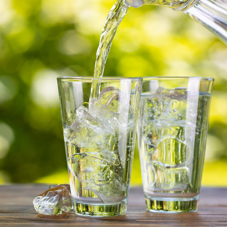 Zwei Gläser stehen auf einem Tisch, beide sind mit Eiswürfeln gefüllt, in ein Glas wird Wasser eingeschenkt.