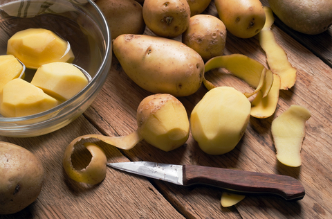 Foto von geschälten Kartoffel in einer Schale mit Wasser, auf einem Holzbrett liegen ungeschälte Kartoffeln und eine halbgeschälte Kartoffel neben einem Messer